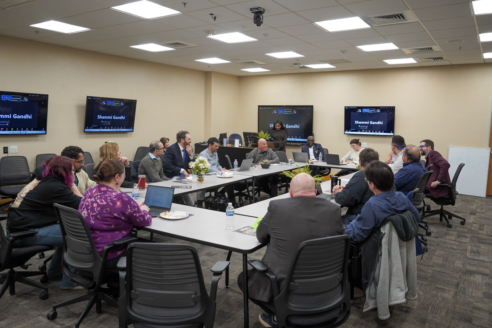 A group of people seated around a conference table in a meeting room, with multiple screens in the background.