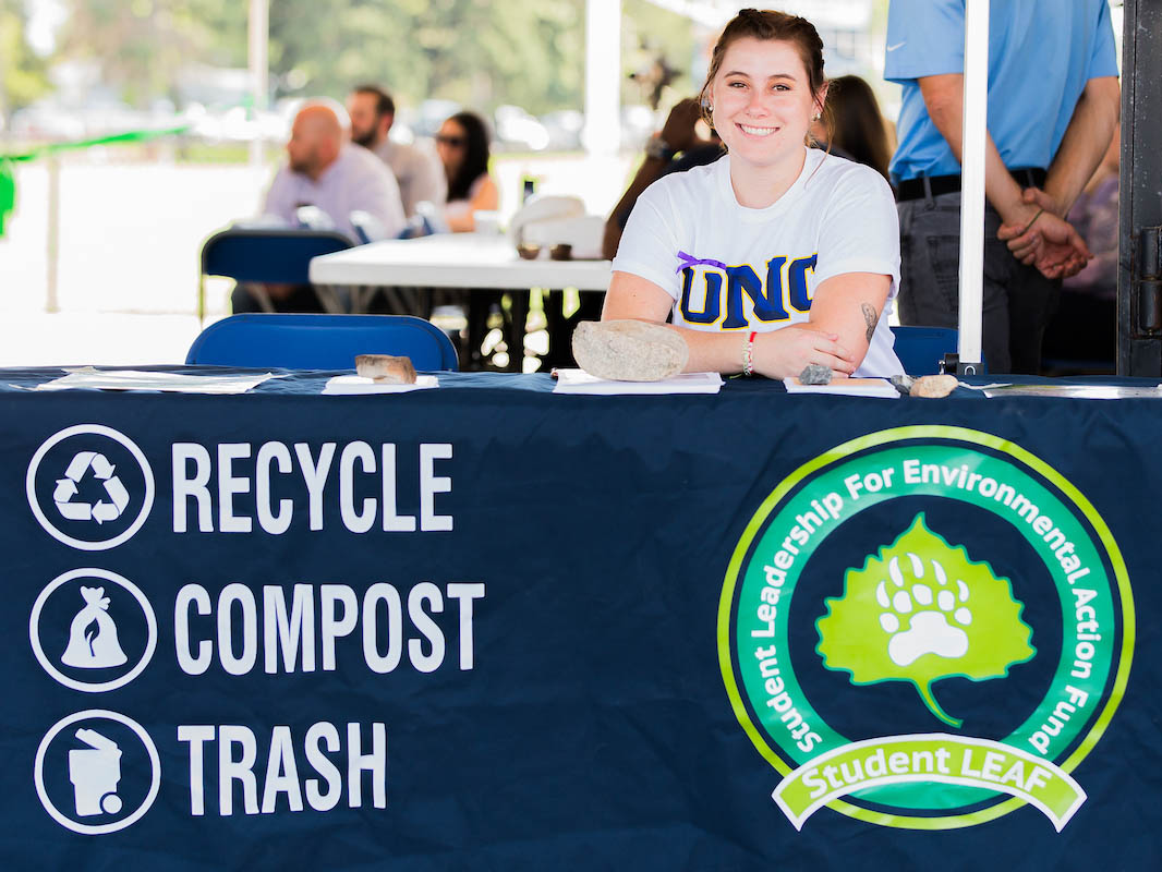 Student sitting behind a table with the Student Leaf logo and recycle. compost, trash.