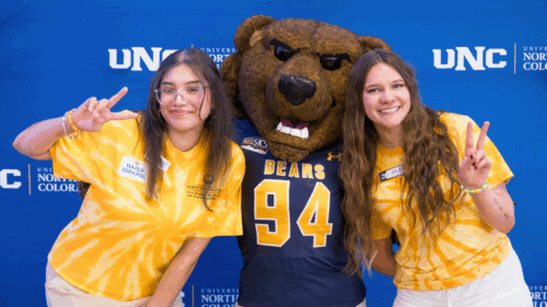 Two student staff members wearing yellow tie-dye UNC shirts pose with the Klawz bear mascot in front of a blue UNC backdrop.