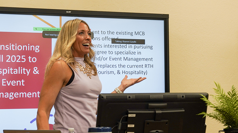 A woman enthusiastically presenting in front of a screen displaying information about transitioning to new hospitality and event management programs. A computer monitor and a plant are visible on the desk beside her.