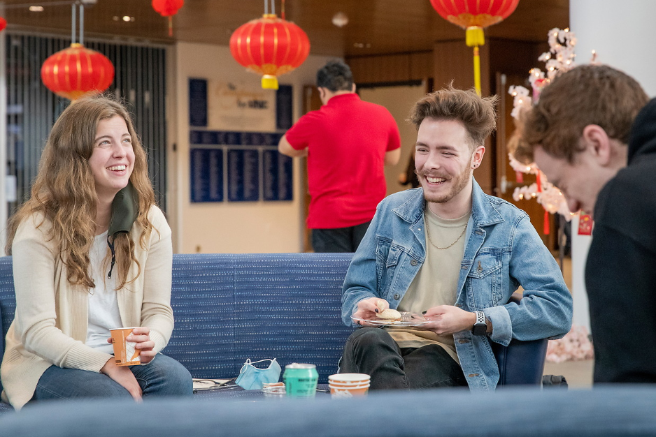 Two students sit on a couch laughing together eating small finger foods.