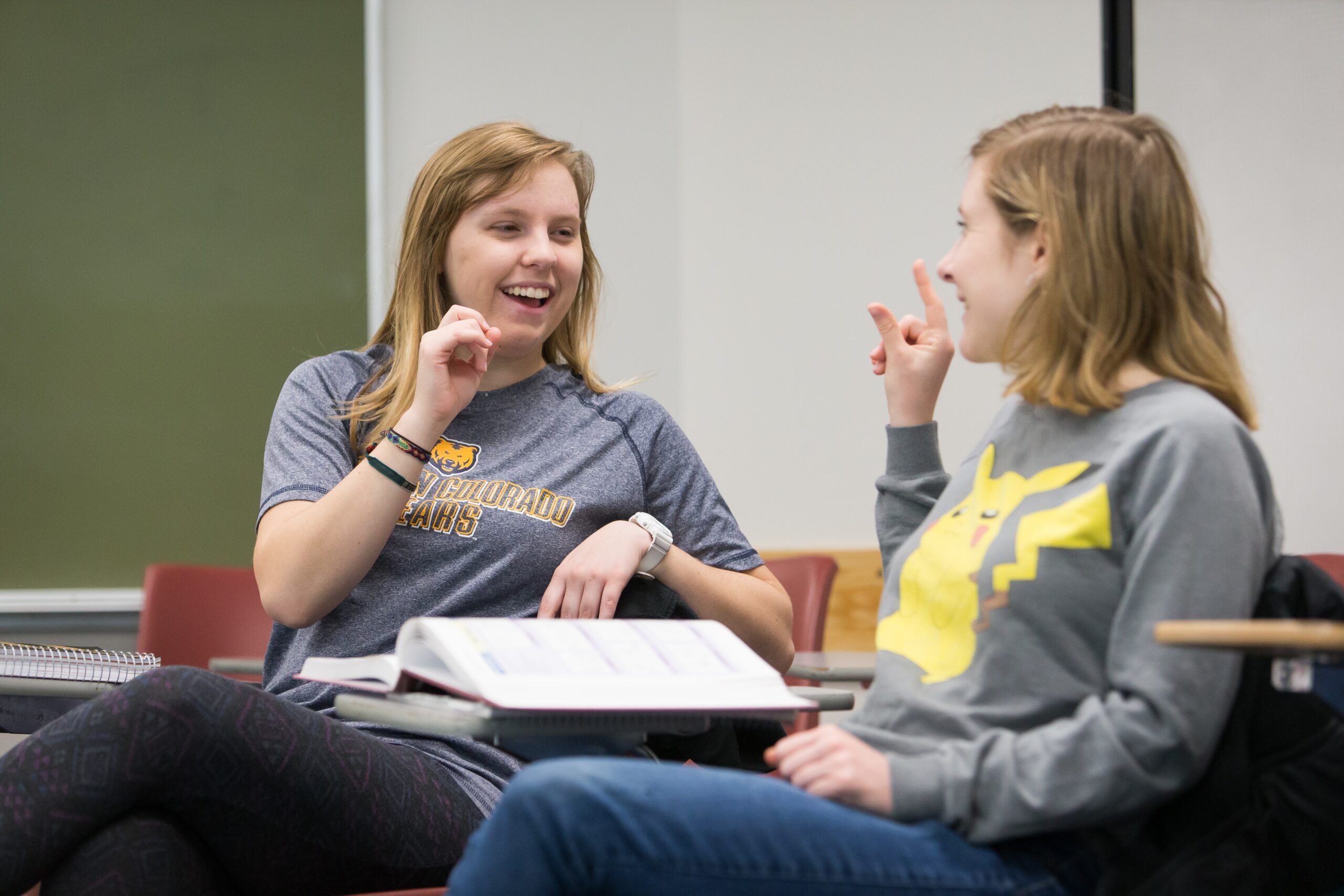 Two college students smiling and practicing American Sign Language together in a classroom.
