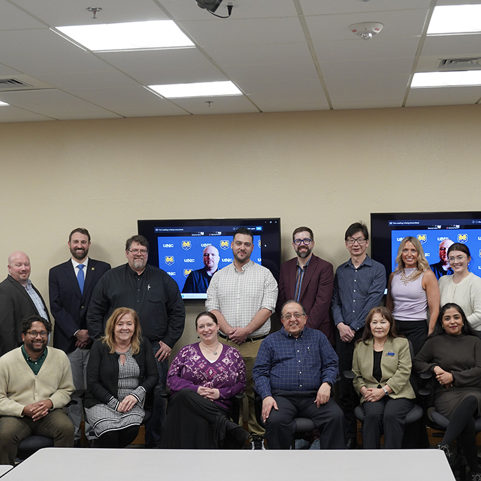 A group of people, standing and sitting, smiling in a brown colored room.