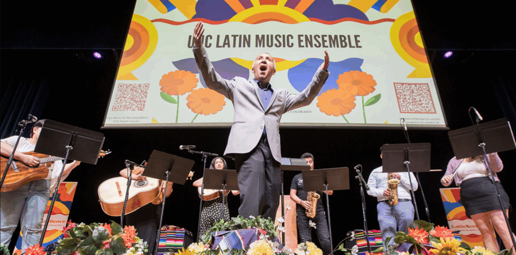 A hispanic man sings on stage while a band plays behind him. A sign behind him reads UNC Latin Music Ensemble.