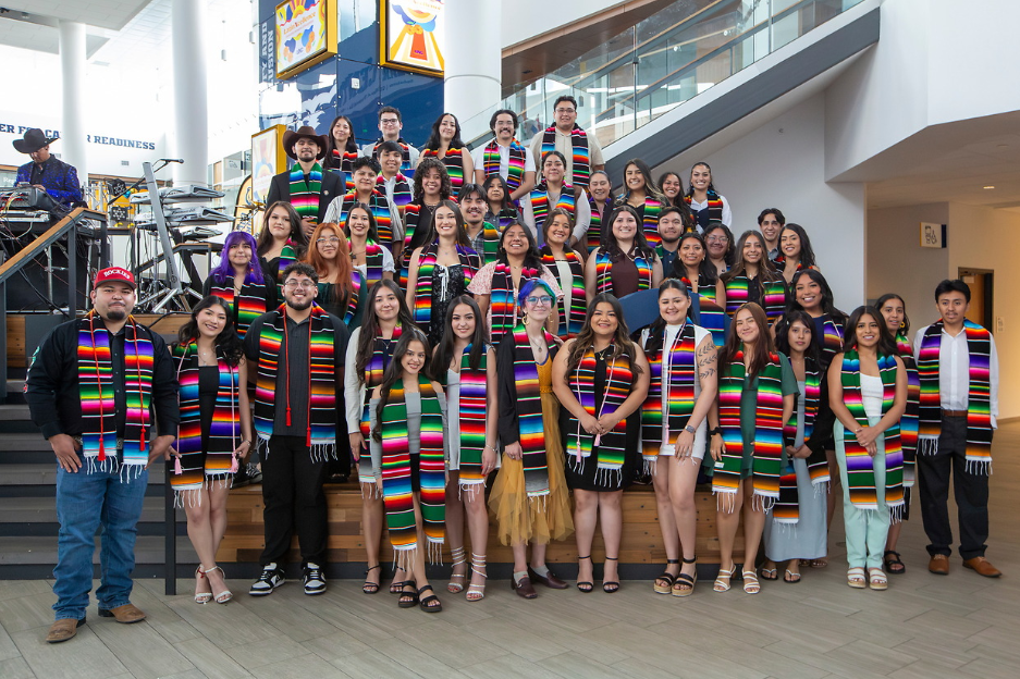 A large group of students stand together on the steps of campus commons wearing coloful scarves.