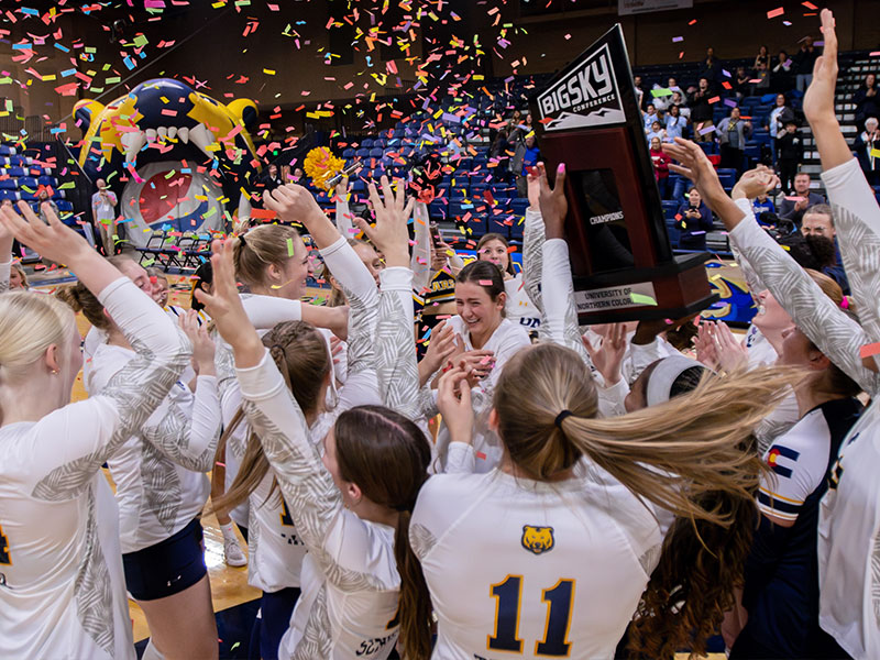 Volleyball celebrates winning the Big Sky Championship by holding up the trophy as confetti falls on them.