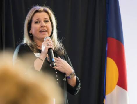 A woman speaking into a microphone in front of the Colorado flag.