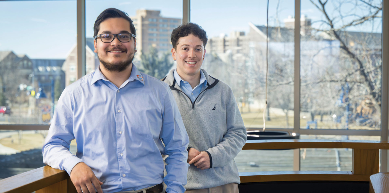 Two students in business-casual clothing smiling at the camera and leaning on a wooden counter.