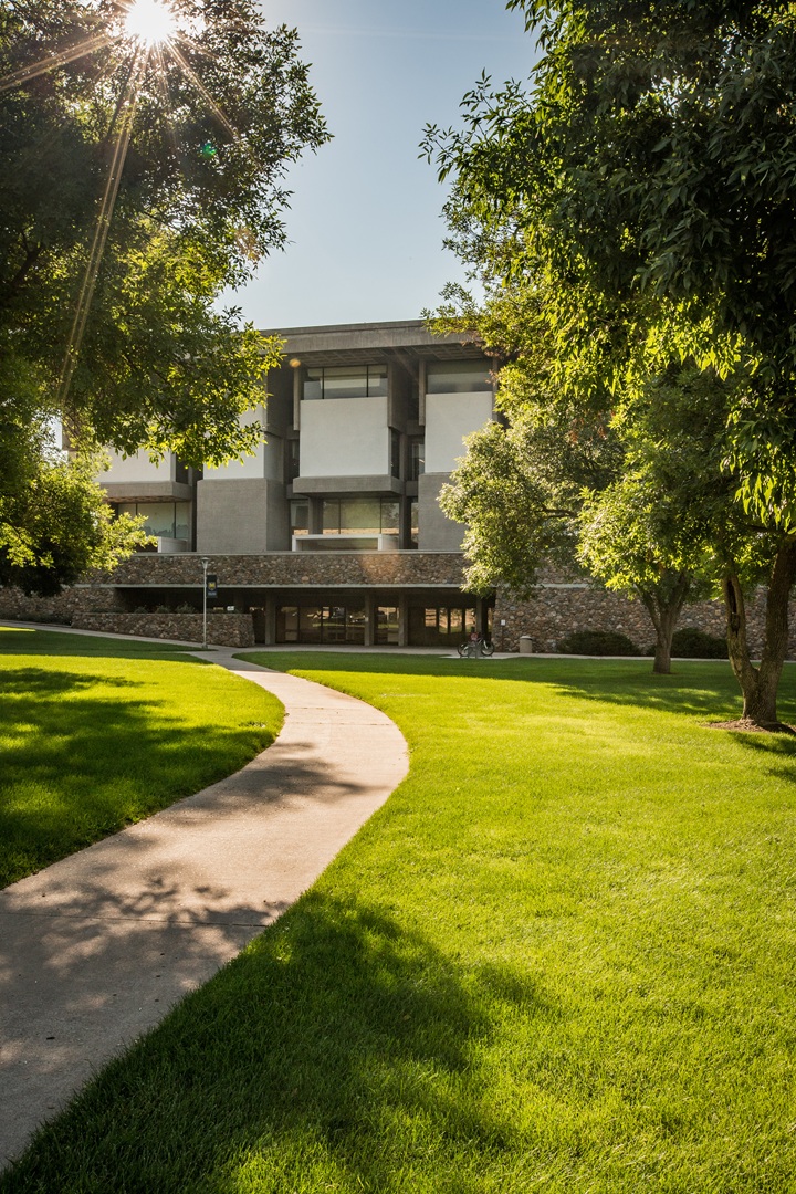 Exterior of Michener Library from a distance, with a path leading to the main entrance. The sun is shining.