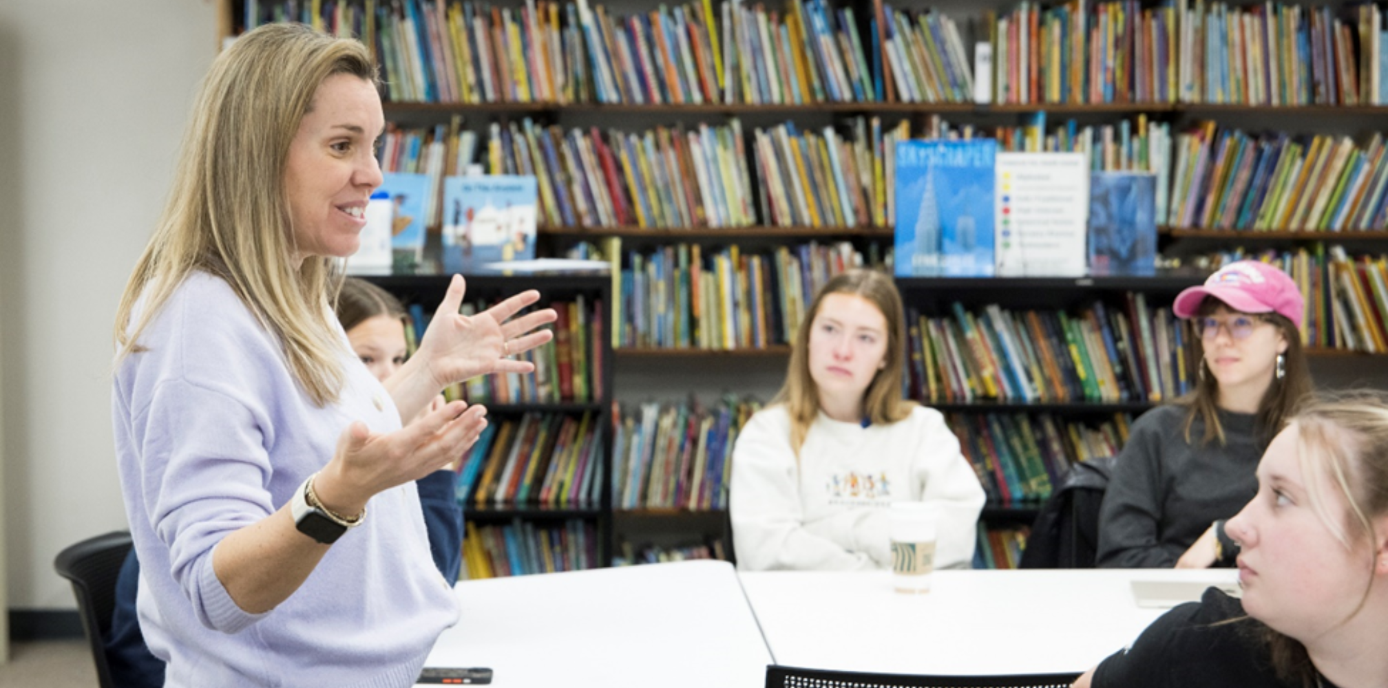 A professor smiling as she speaks to group of students in a library.