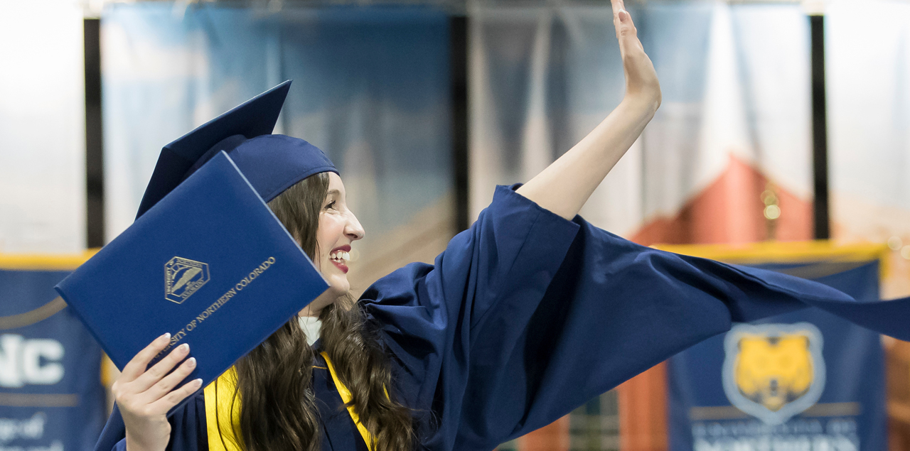 Closeup of graduate holding diploma, smiling and waving to the crowd.