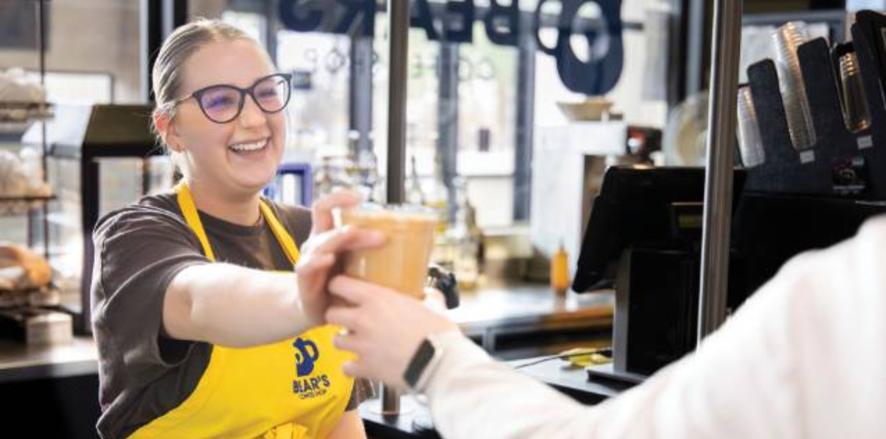 A student in a yellow apron smiling as she hands a cup of coffee to a customer.
