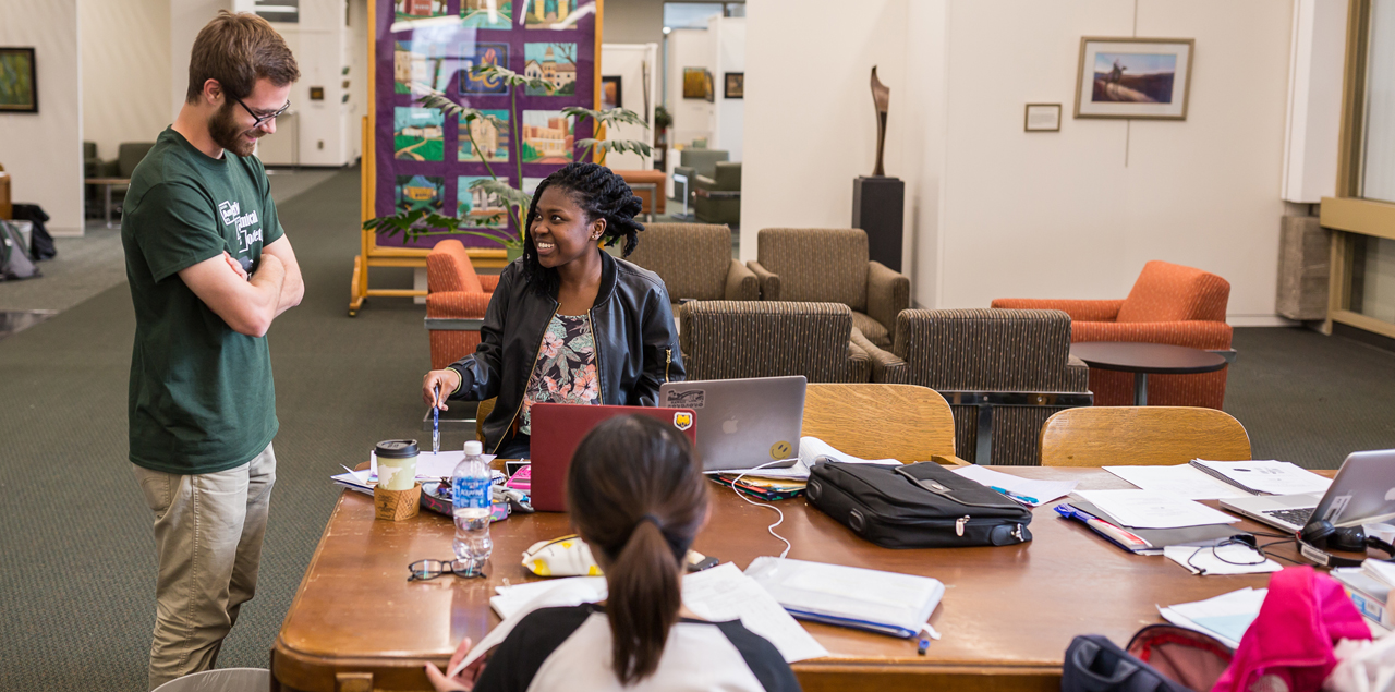Three students at a table in the library with books and papers spread out.
