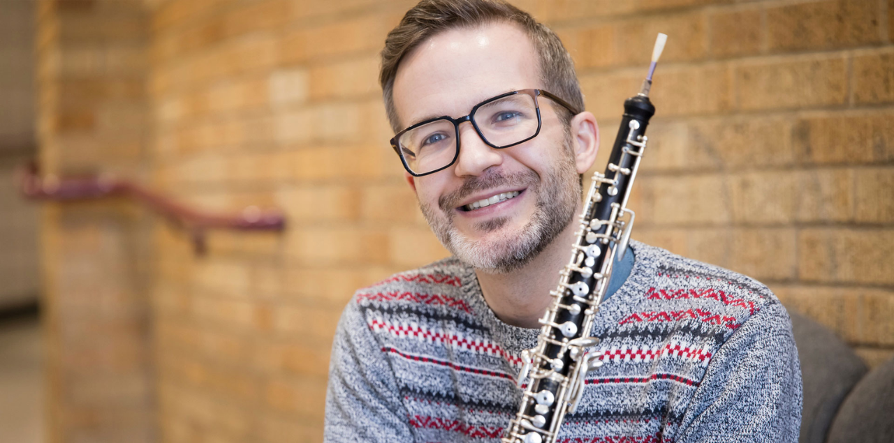 A man smiling and sitting down while holding a woodwind instrument.