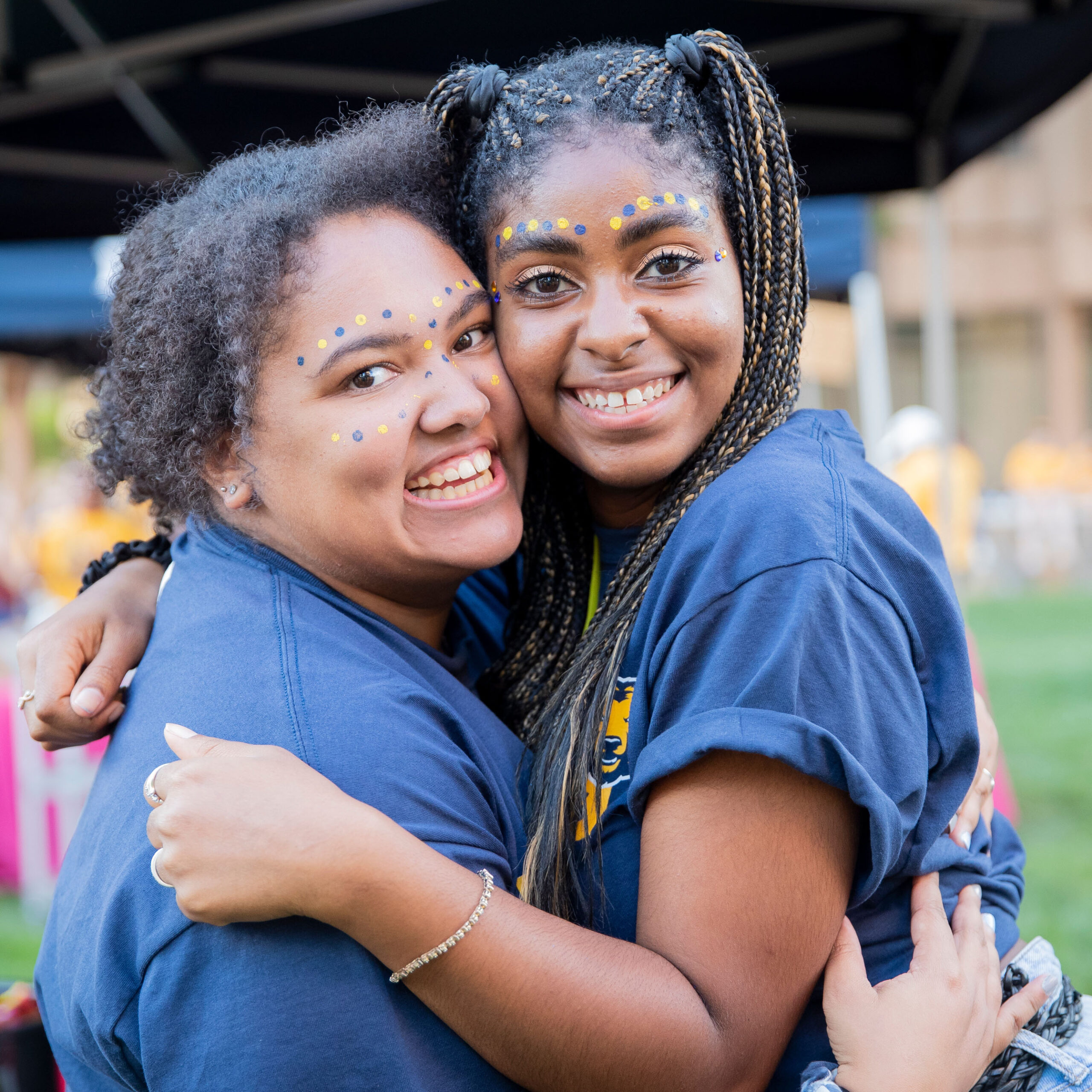 Two young women smiling and hugging, with colorful face paint and casual blue T-shirts, under a canopy.