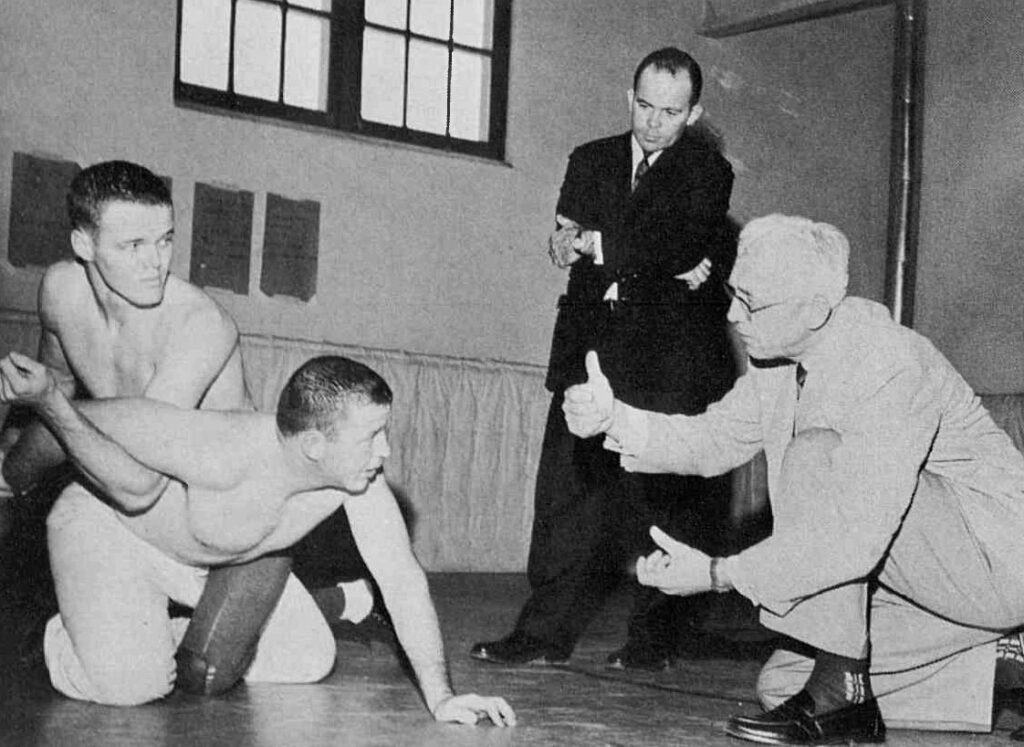 A black-and-white photo of two men wrestling while a coach gives them direction.