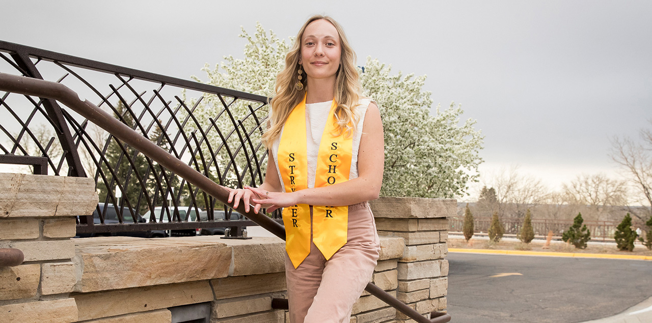 Madeline Dannewitz standing on stairs on campus with her graduation stole around her neck