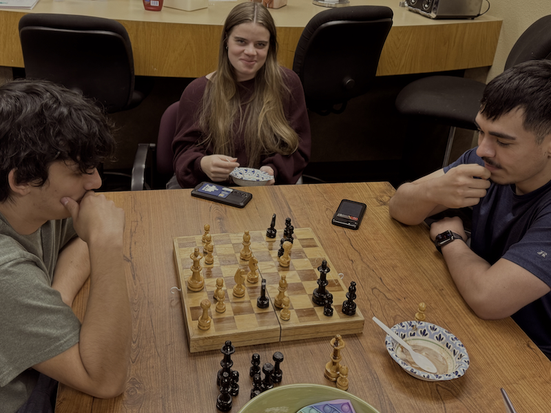 Two male students playing chess while additional student looks on while eating ice cream.