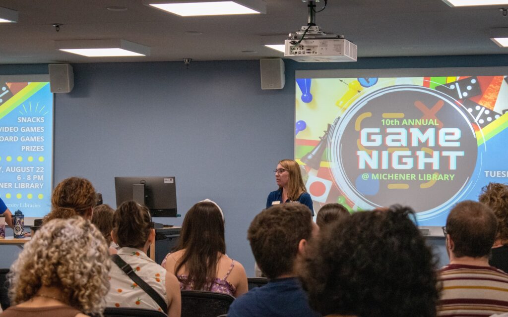 Students attending Welcome Day at Michener Library. Students are seated and listening to a library employee speak. There is a slide about Game Night on the projector behind the speaker.