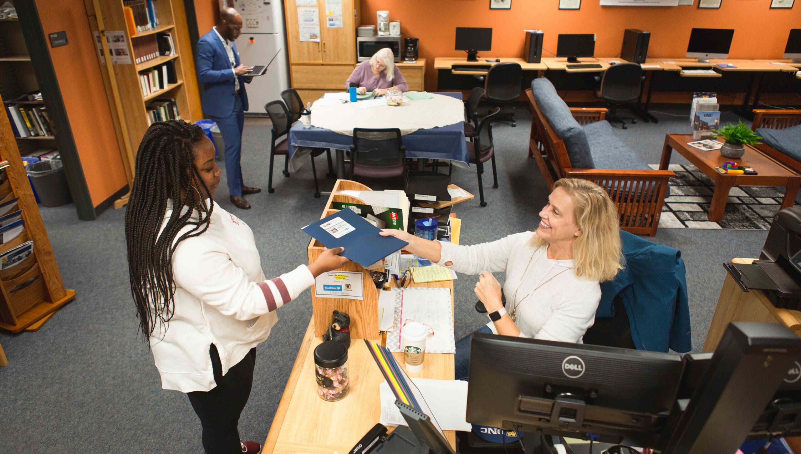 A woman at a library or office desk hands a folder to another woman standing in front of her. Computers and bookshelves are visible in the background, with a few other people working in the area. The setting is bright and organized.