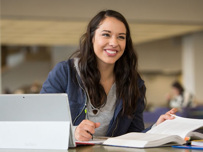 A student smiles while taking notes and working on her laptop.