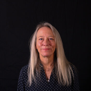 A woman smiling, dressed in a black shirt, against a black background.
