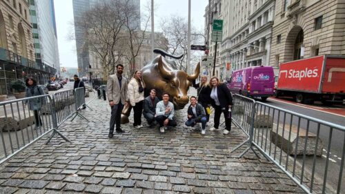 A group of FMA students posing in front of the Charging Bull statue on a cobblestone street in New York City, with surrounding buildings and a Staples truck in the background.