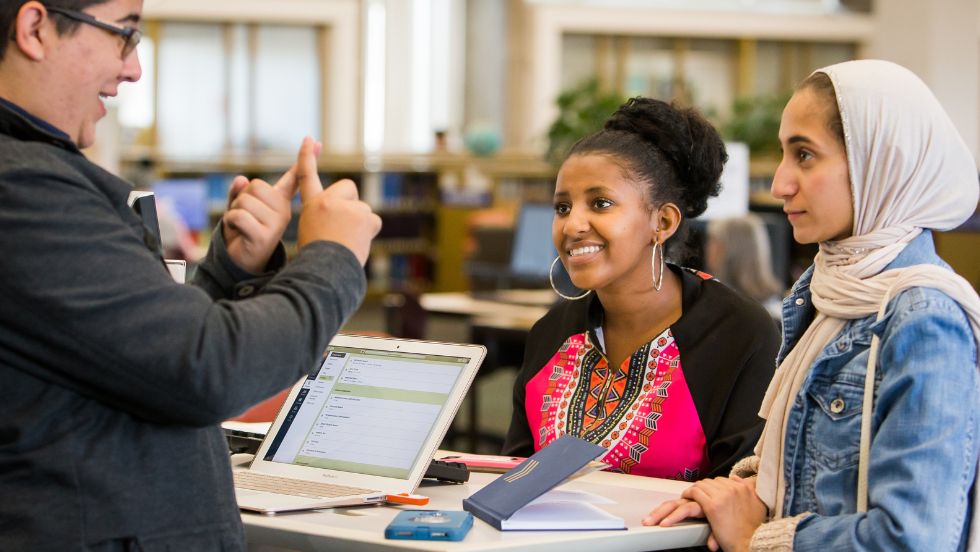 Two students speak to a friendly person at a service desk.
