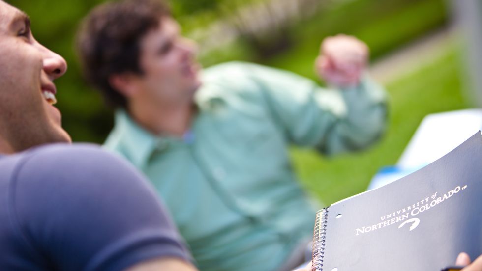 A student holds a notebook and smiles as he engages with others on campus.