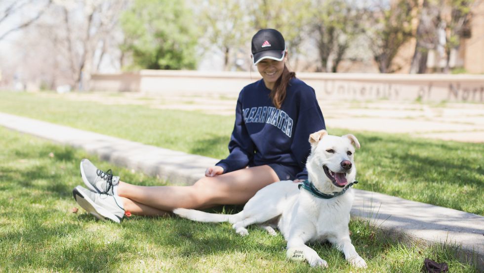 A dog sits with their owner on the grass on campus