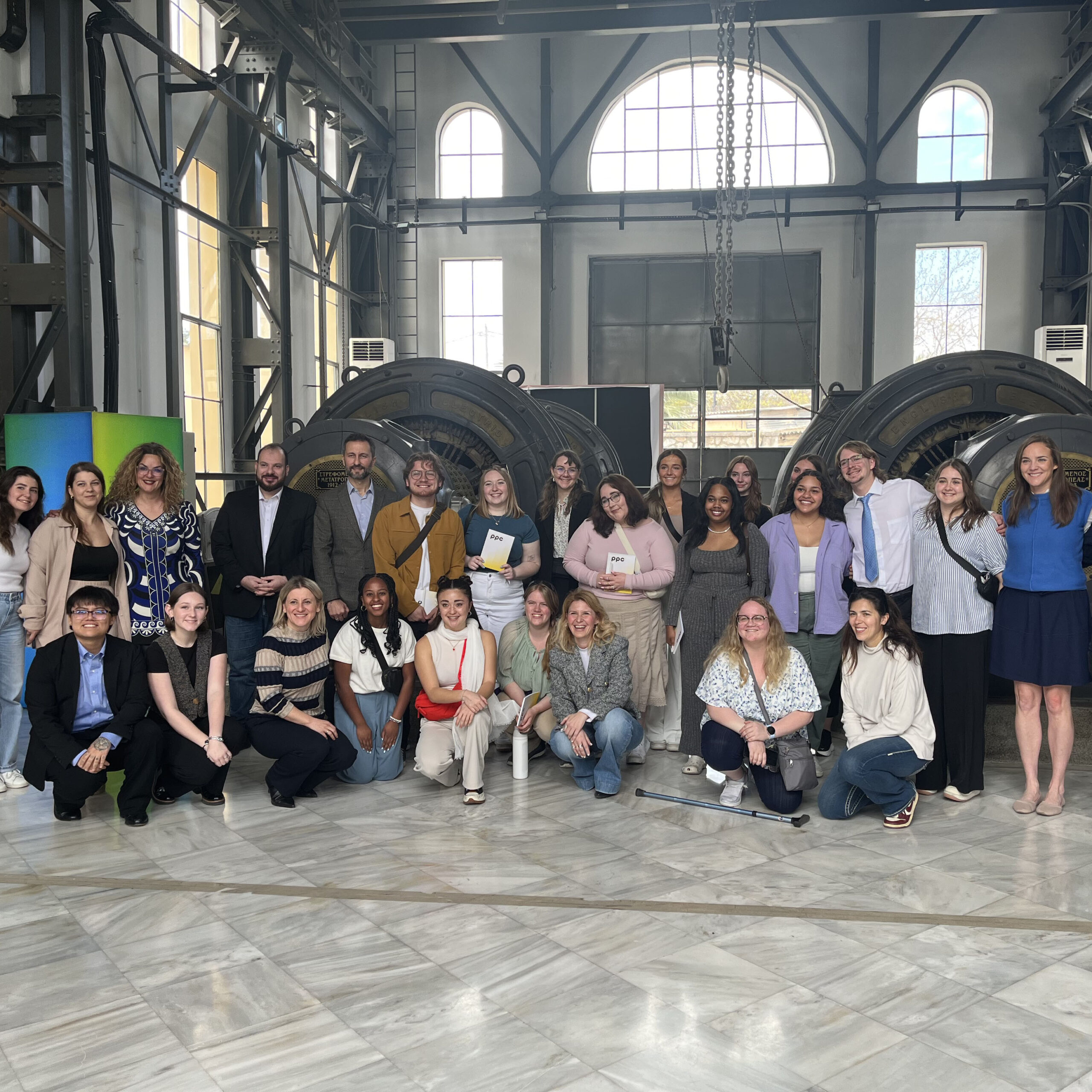 A group of diverse young professionals and students gathered together indoors in a spacious industrial-style building with large windows and machinery in the background.