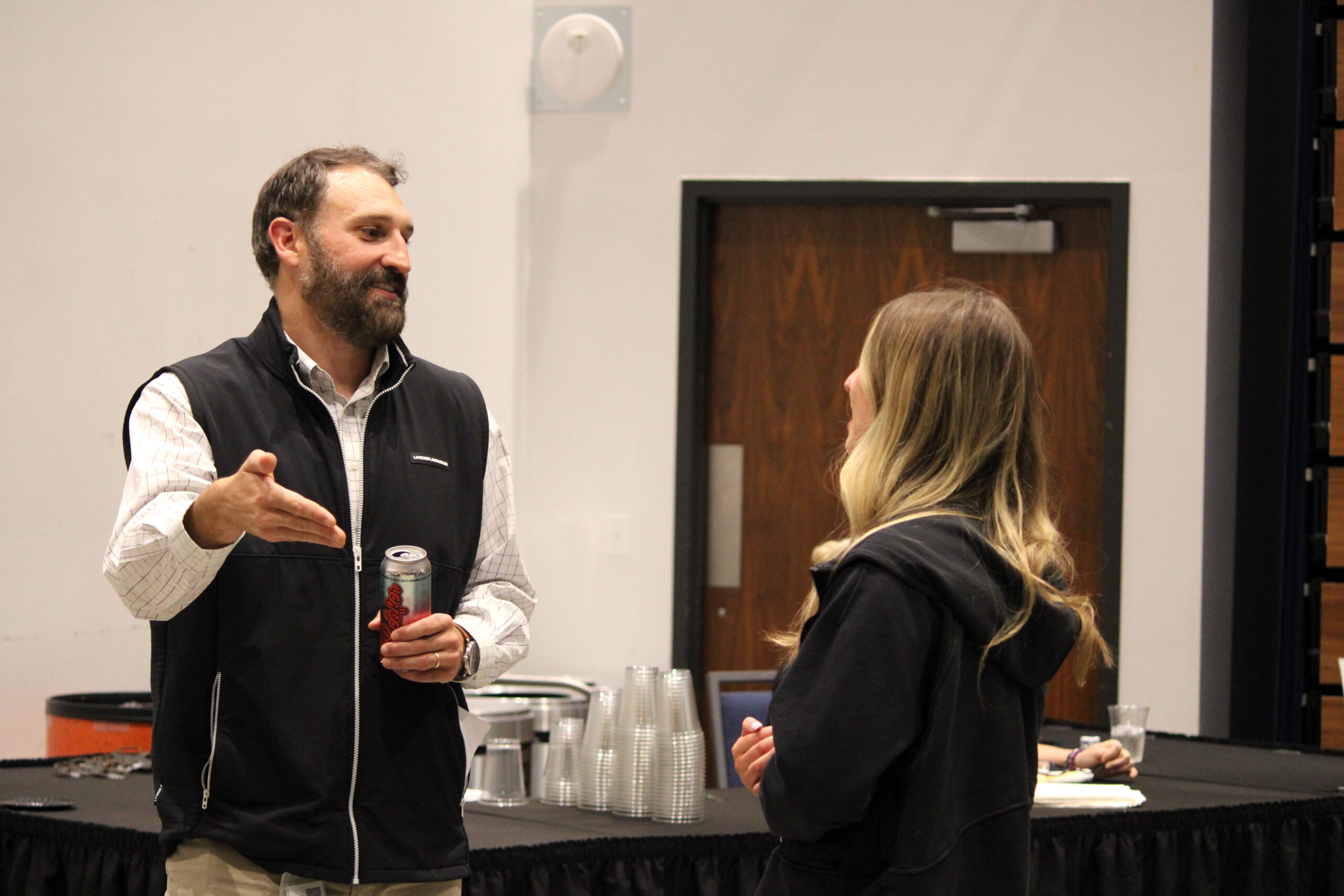 A man and a woman are conversing in a room with a white wall and a wooden door. The man, wearing a black vest, holds a drink can and gestures with his hand. The woman is facing him, wearing a dark jacket.