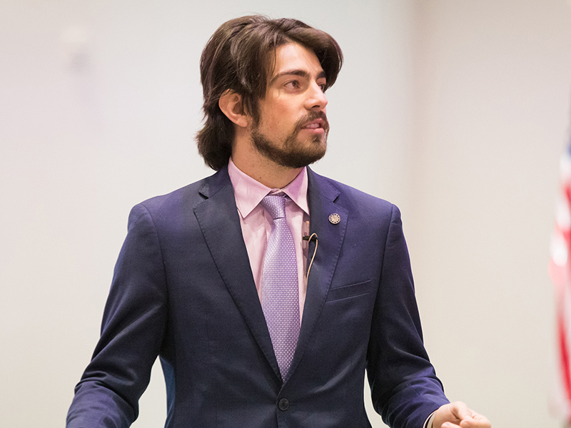 A male student with dark hair and light skin, presenting, dressed in business professional attire.