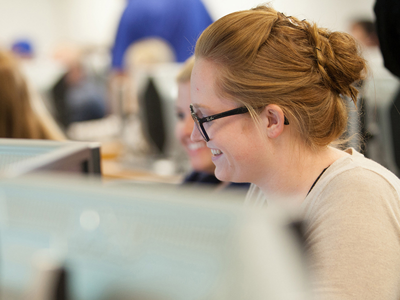 A smiling young woman with red hair tied back and wearing glasses, working at a computer in a busy classroom or computer lab setting.
