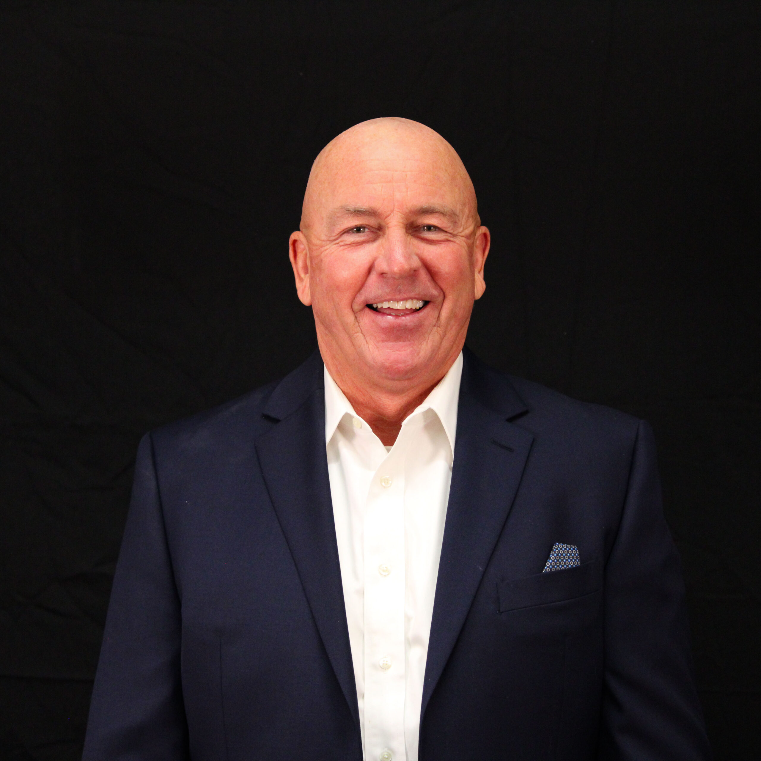 A person smiling, dressed in a dark suit with a white shirt and a pocket square, against a black background.