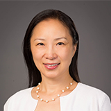 A woman smiling, dressed in a white shirt with a pearl necklace, against a grey background.