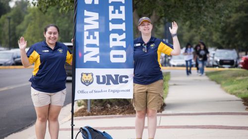 Two orientation leaders in navy and gold UNC shirts stand on the sidewalk waving next to a tall banner that reads “New Student Orientation.”