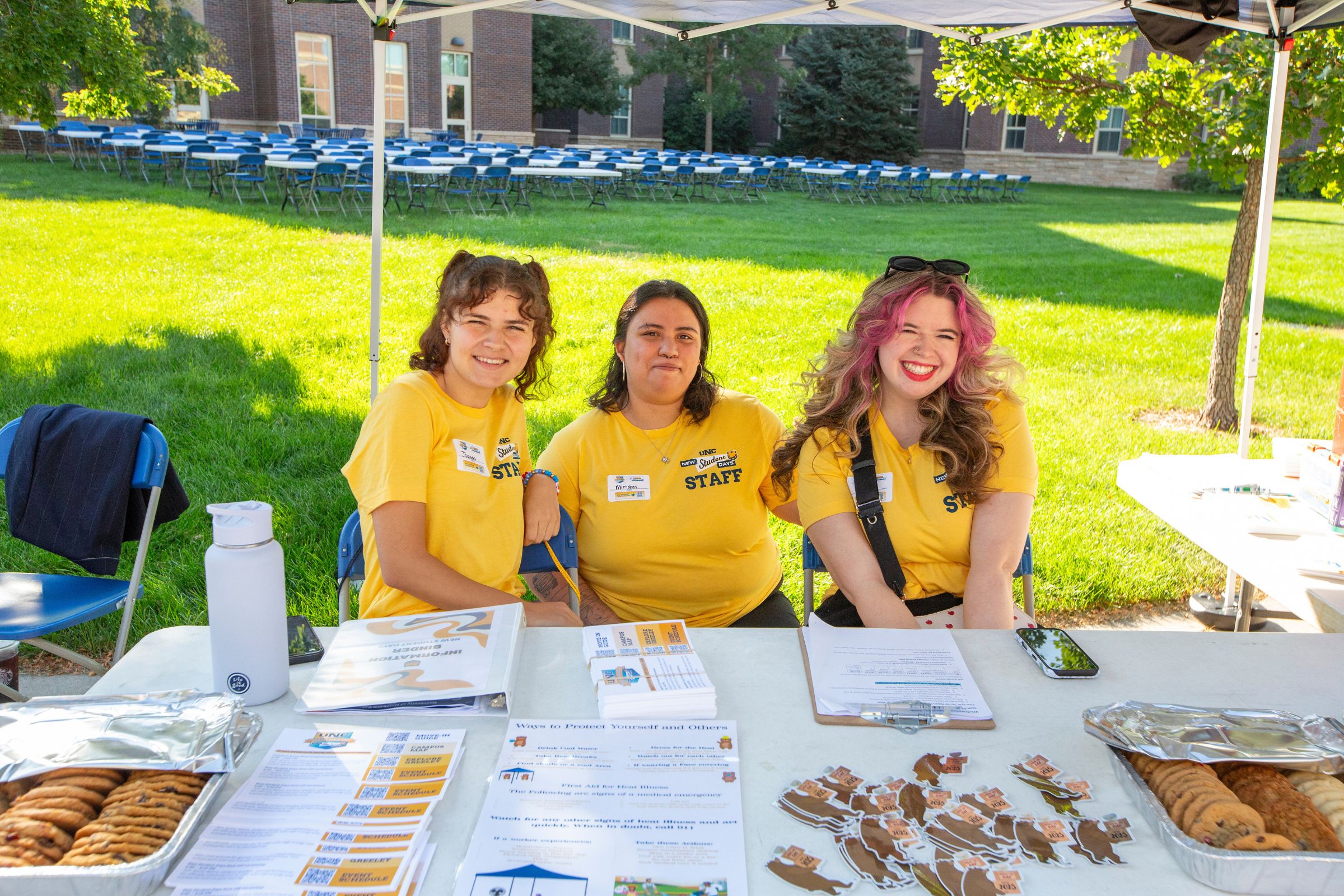 Three UNC student staff members in yellow tie-dye shirts sit at an outdoor check-in table. They smile at the camera, surrounded by informational handouts, stickers, and trays of cookies.