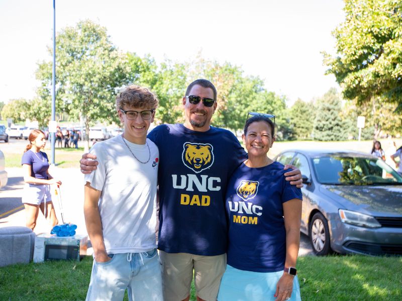A smiling student stands with their parents during UNC Move-In Day. The parents wear matching navy shirts that read “UNC Dad” and “UNC Mom,” while the student stands between them in a white T-shirt and jeans.