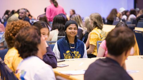 Students sit around round tables in a large meeting room, engaging in small group discussions during orientation.