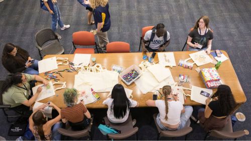 Students sit around a large table indoors decorating canvas tote bags with colorful markers while others stand nearby chatting