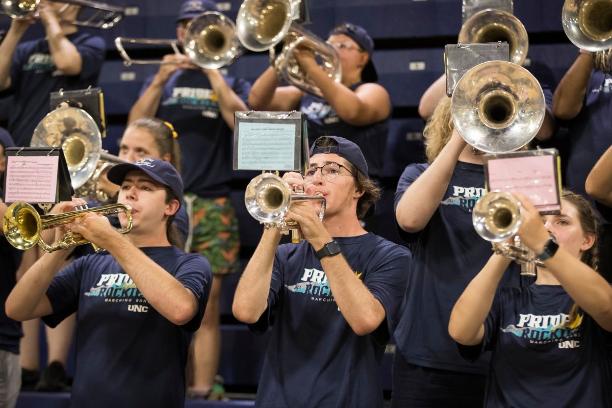 UNC marching band members play brass instruments indoors, wearing navy “Pride of the Rockies” shirts and ball caps.