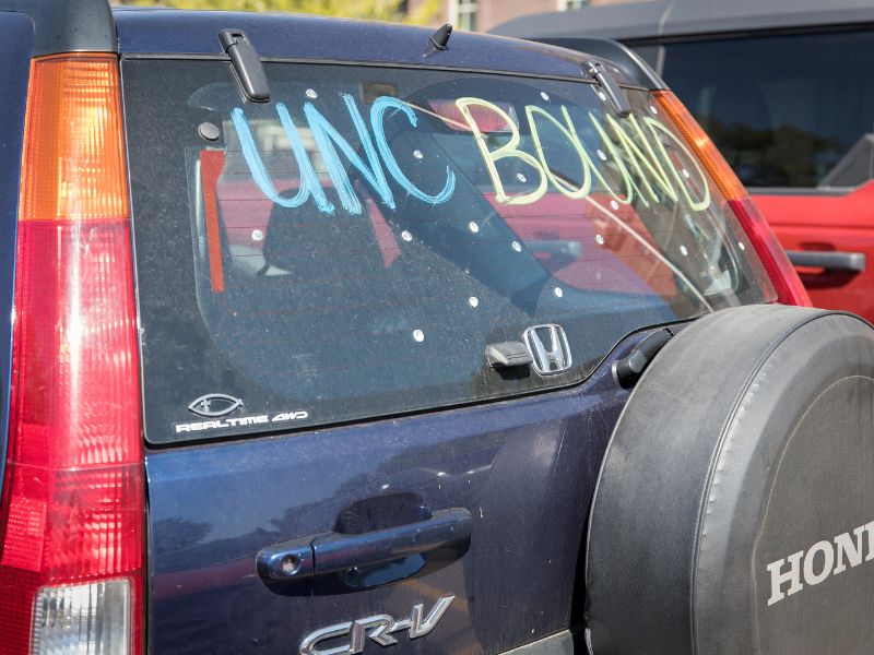 The back window of a blue Honda CR-V decorated with colorful paint reads “UNC Bound,” parked among other cars.