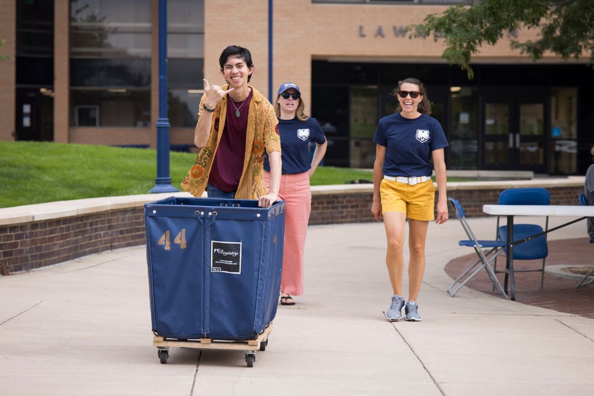 A smiling student pushes a large blue move-in cart outside a residence hall while two UNC staff members in matching shirts walk alongside, laughing and supporting.