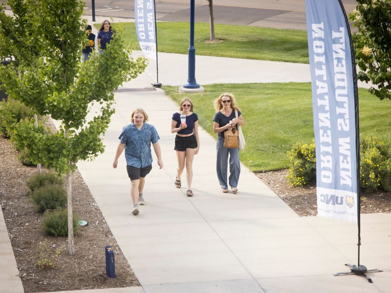 Students and family members walk up a campus sidewalk toward New Student Orientation banners, carrying belongings and drinks.