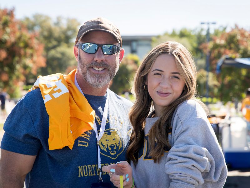 A father and daughter pose together at a UNC homecoming tailgate. The father wears a blue UNC Bears shirt and sunglasses with a yellow T-shirt draped over his shoulder, while the daughter smiles in a gray UNC sweatshirt.