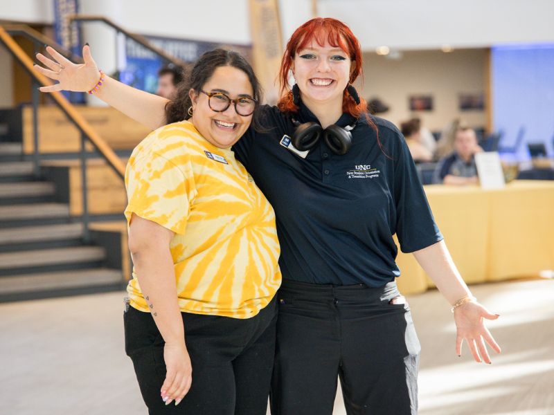Two smiling student staff members, one in a yellow tie-dye UNC shirt and the other in a navy polo, pose with arms around each other inside the check-in area.