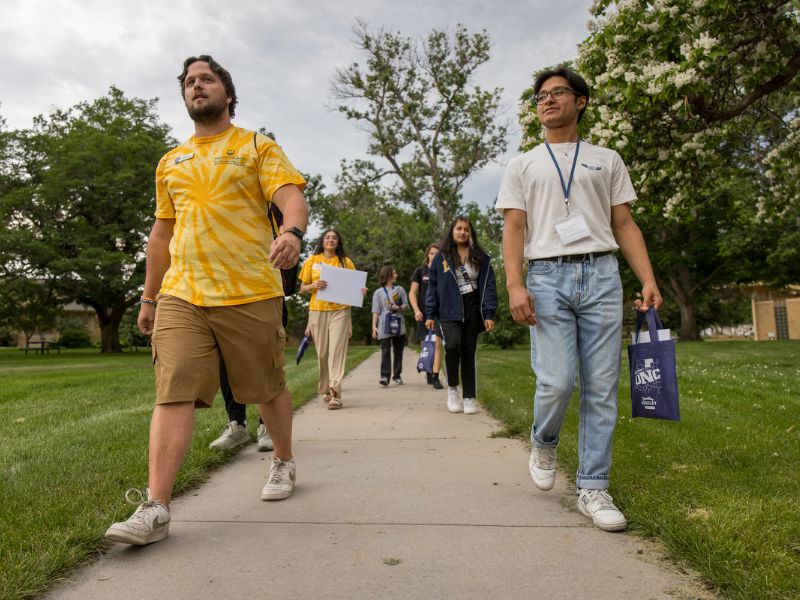 A group of orientation leaders and students walk across campus sidewalks in pairs, carrying UNC tote bags and papers.