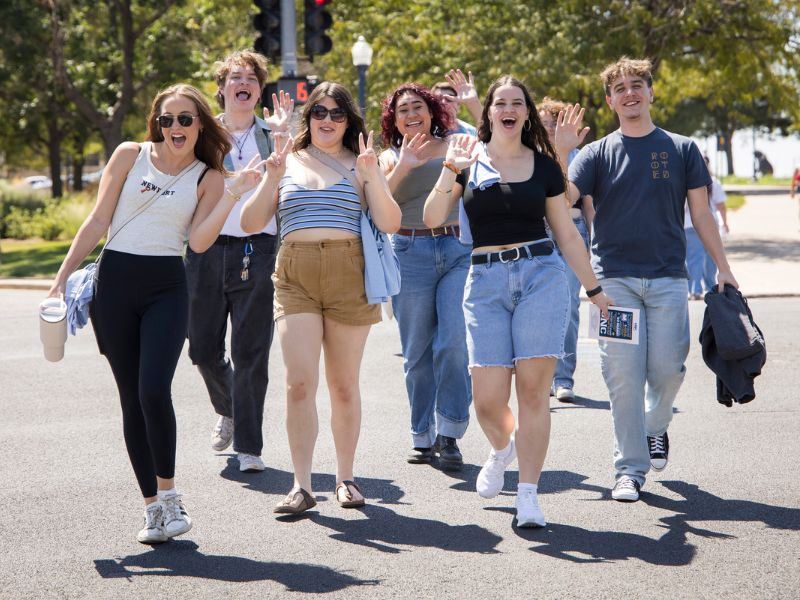 A group of friends smile and wave enthusiastically while crossing a campus street together on a sunny day.