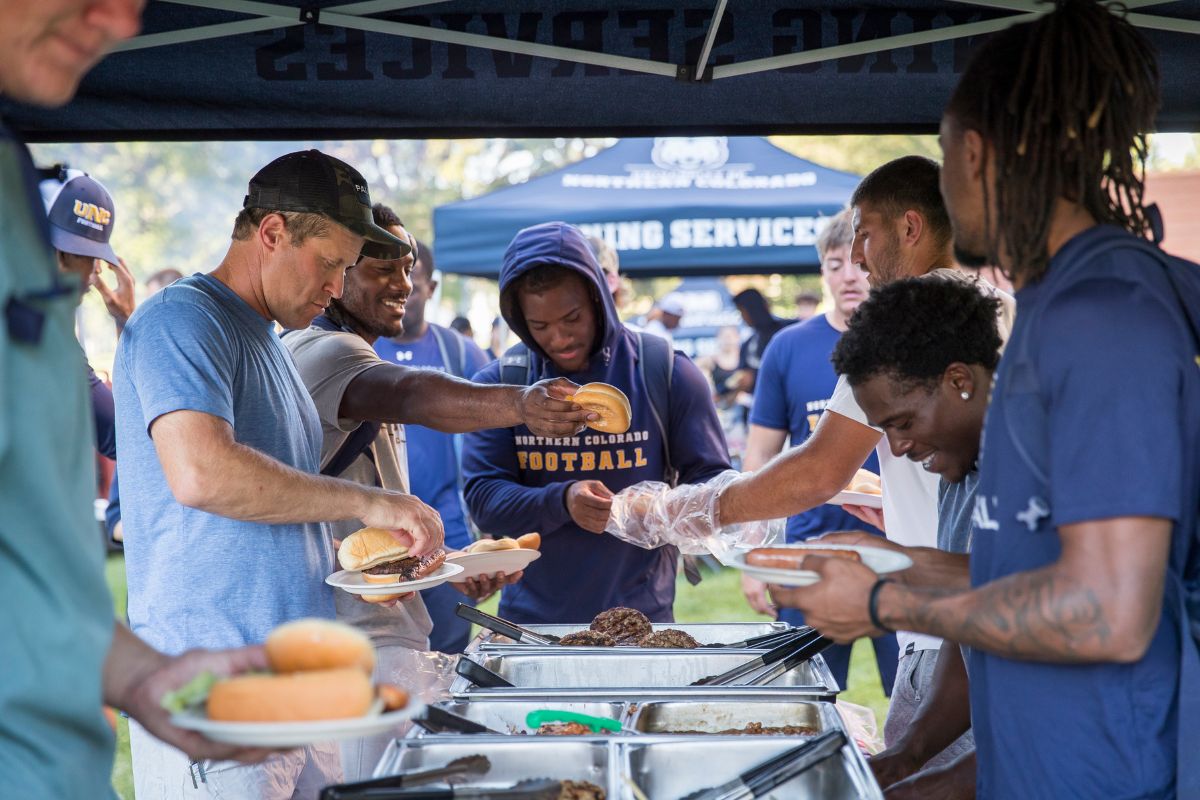 Students and staff line up under a UNC Dining Services tent serving hamburgers and food from large trays during an outdoor welcome event.